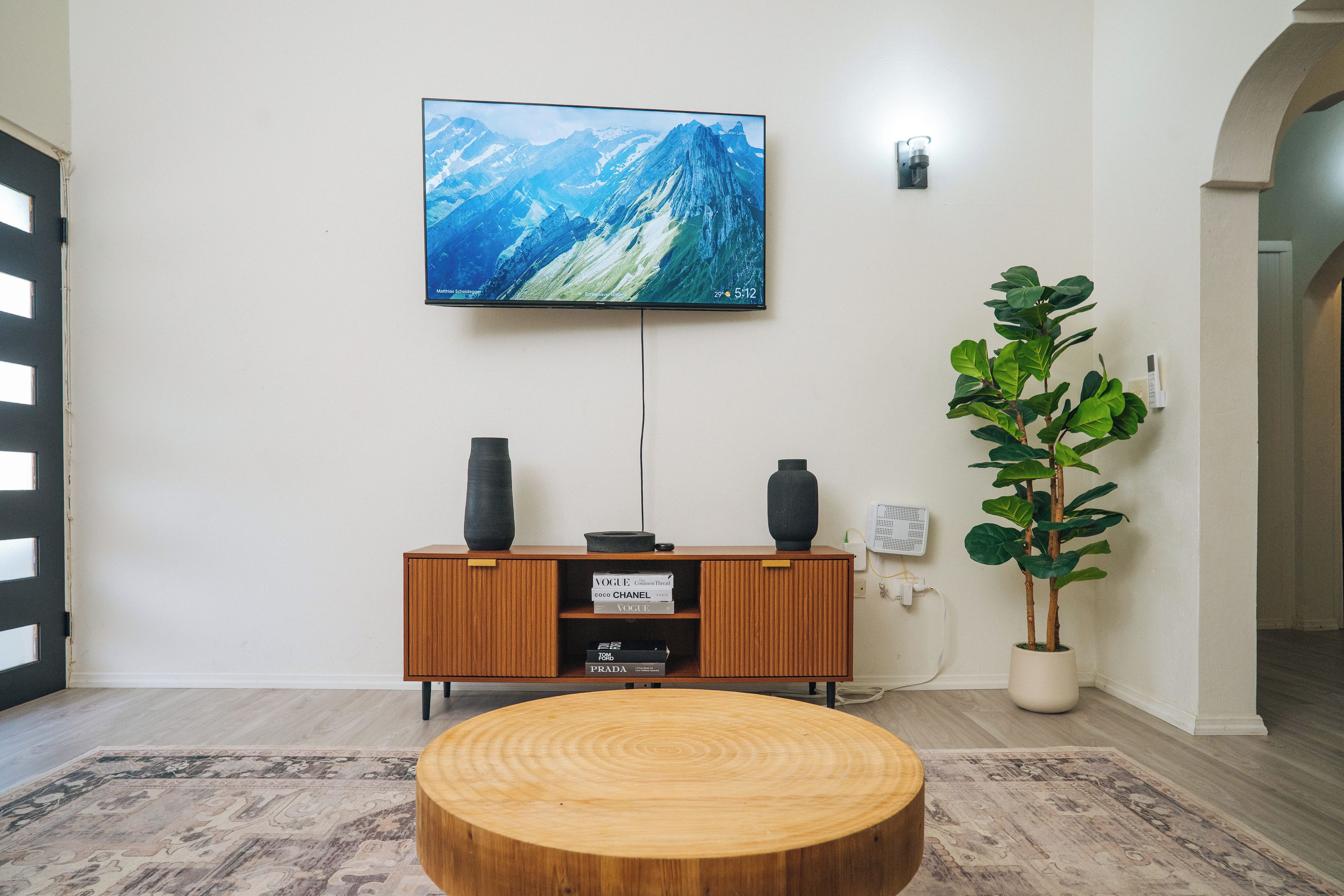 A living room with a flat-screen TV mounted on the wall, a wooden cabinet with books, a round wooden coffee table, and a potted plant.