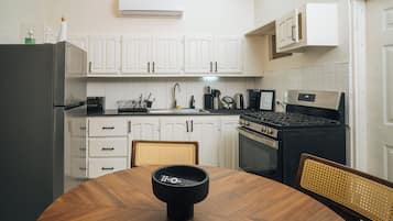 A kitchen with white cabinets, a black stove, a dining table with chairs, and an air conditioning unit above the cabinets.
