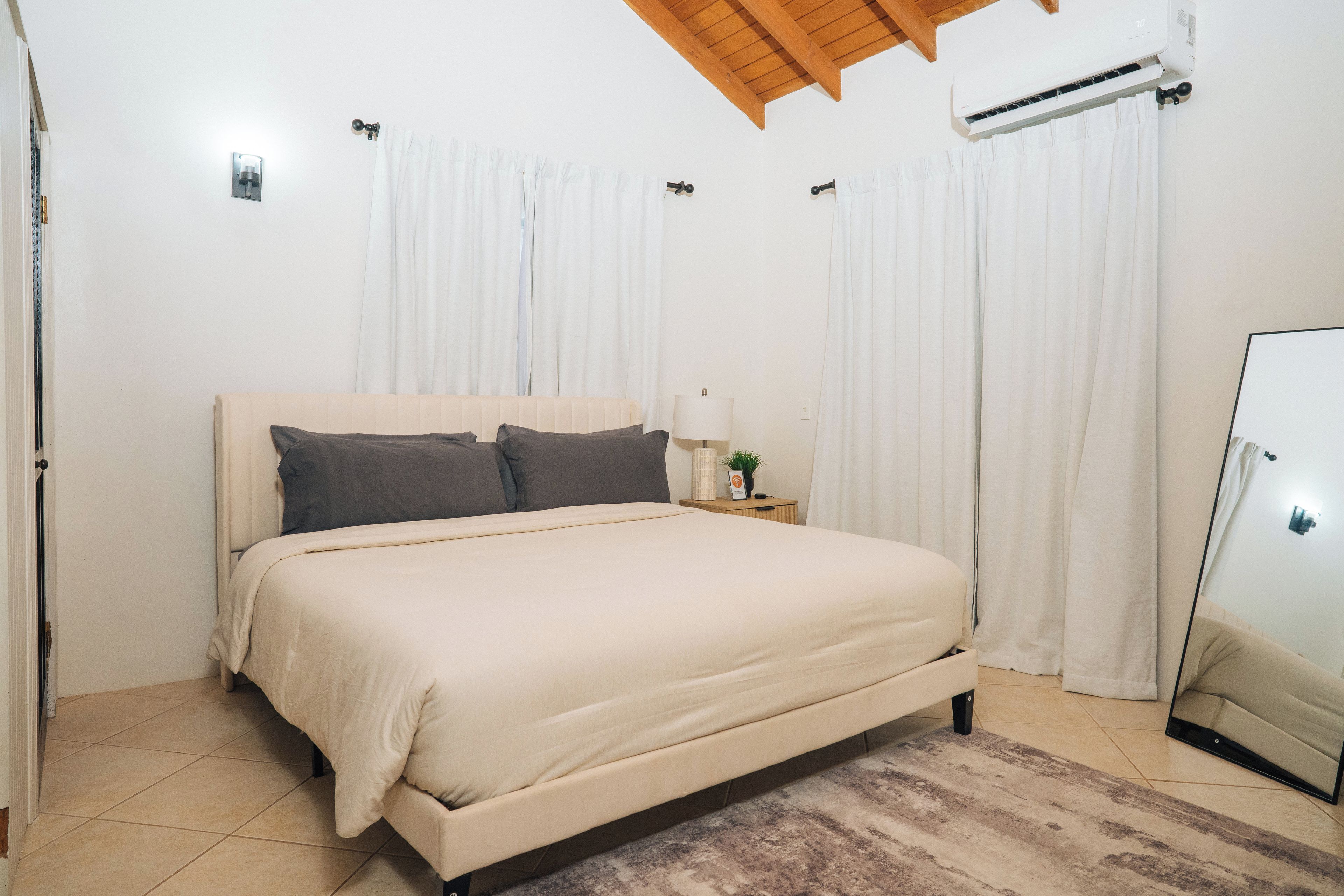 Ein Schlafzimmer mit Bett, Vorhängen, einem Nachttisch, einer Lampe und einer Klimaanlage.