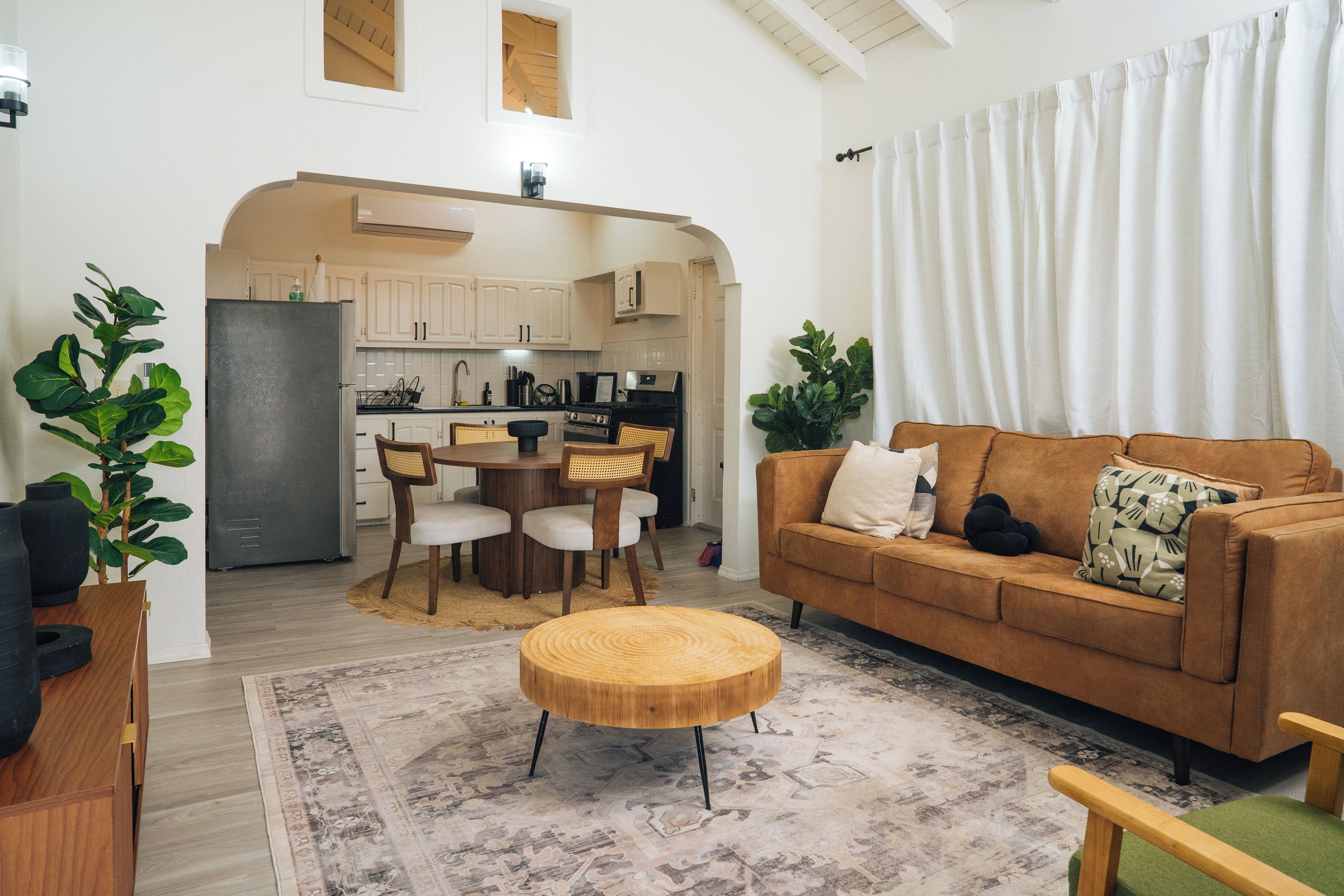 Ein modernes Wohnzimmer mit einem braunen Sofa, einem runden Holztisch und einem Essbereich mit weißem Tisch und Stühlen.