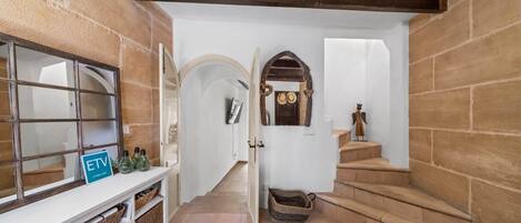 Entrance hall with sandstone walls, exposed wooden beams and a console with wicker baskets, giving access to an arched hallway and the staircase leading to the upper floor.