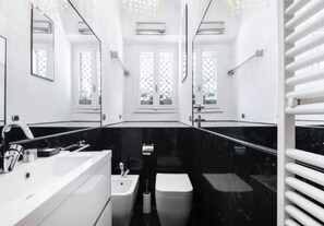 A modern bathroom with a white vanity, a sink, a toilet, and a bidet. There are mirrors on the walls, and the floor is black marble.