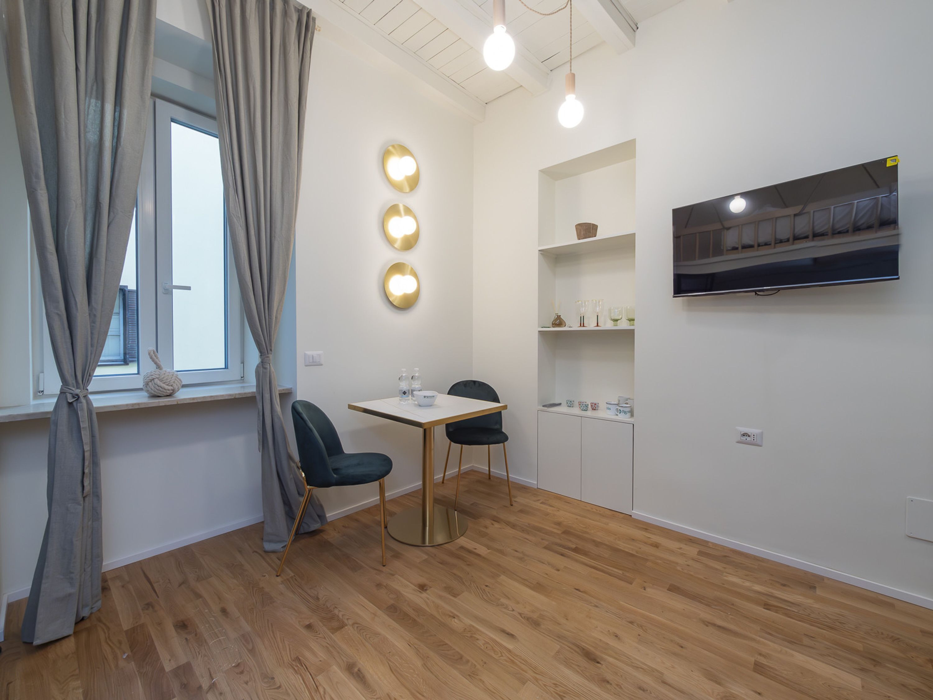 Una habitación moderna y bien iluminada con una mesa de comedor, dos sillas, un televisor montado en la pared y una ventana con cortinas.