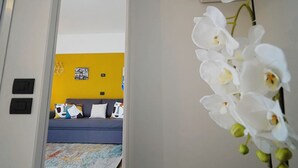 Een deuropening naar een kamer met een bank, kleurrijke kussens en een gele muur met een poster.