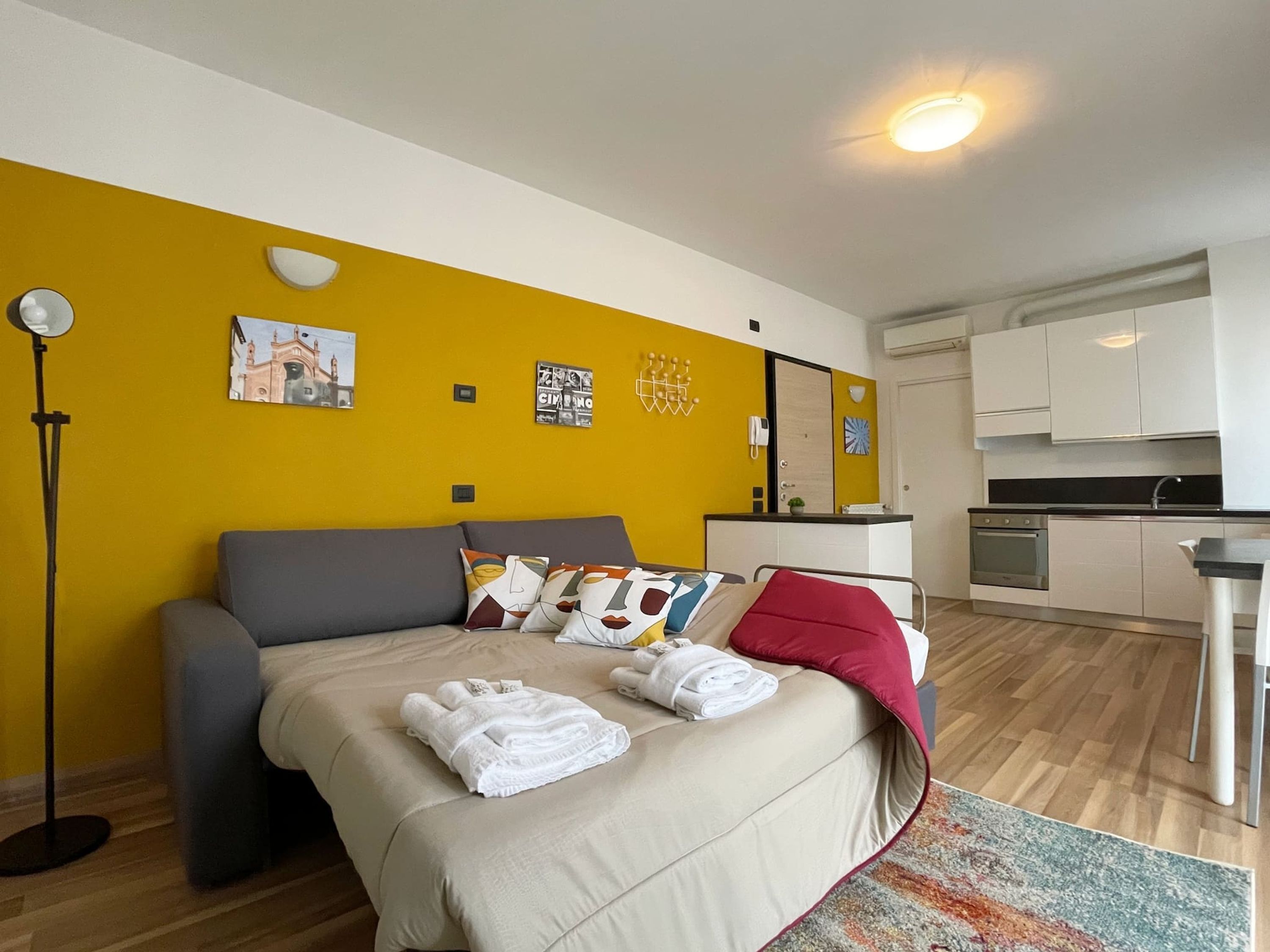 A modern hotel room with a bed, a sofa, a small kitchen area, and a bright yellow accent wall.