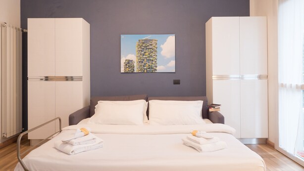 Una habitación de hotel moderna con una cama, dos armarios blancos, una ventana con persianas y un cuadro de paisaje urbano en la pared.