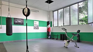 Un gimnasio con paredes blanco y verde, un saco de boxeo, una bicicleta estática y una ventana con persianas.