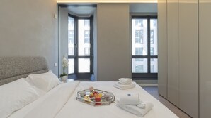 A hotel room with a neatly made bed, a tray of fruit and a rose, and a view of the city through large windows. - Central Tower S Babila - Elegant 2bdr With Rooftop Terrace (Milan)