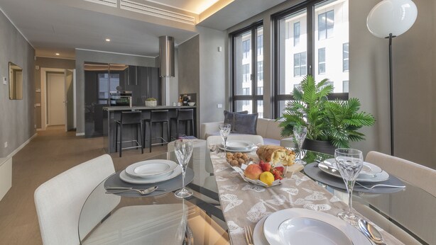 A dining area with a table set for four, a kitchen in the background, and large windows with a view of buildings. - Central Tower S Babila - Elegant 2bdr With Rooftop Terrace (Milan)