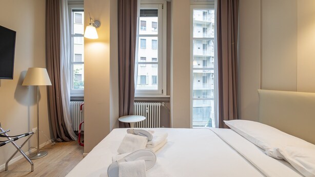 Ein Hotelzimmer mit Bett, kleinem Tisch, Lampe und Blick auf Gebäude durch die Fenster.
