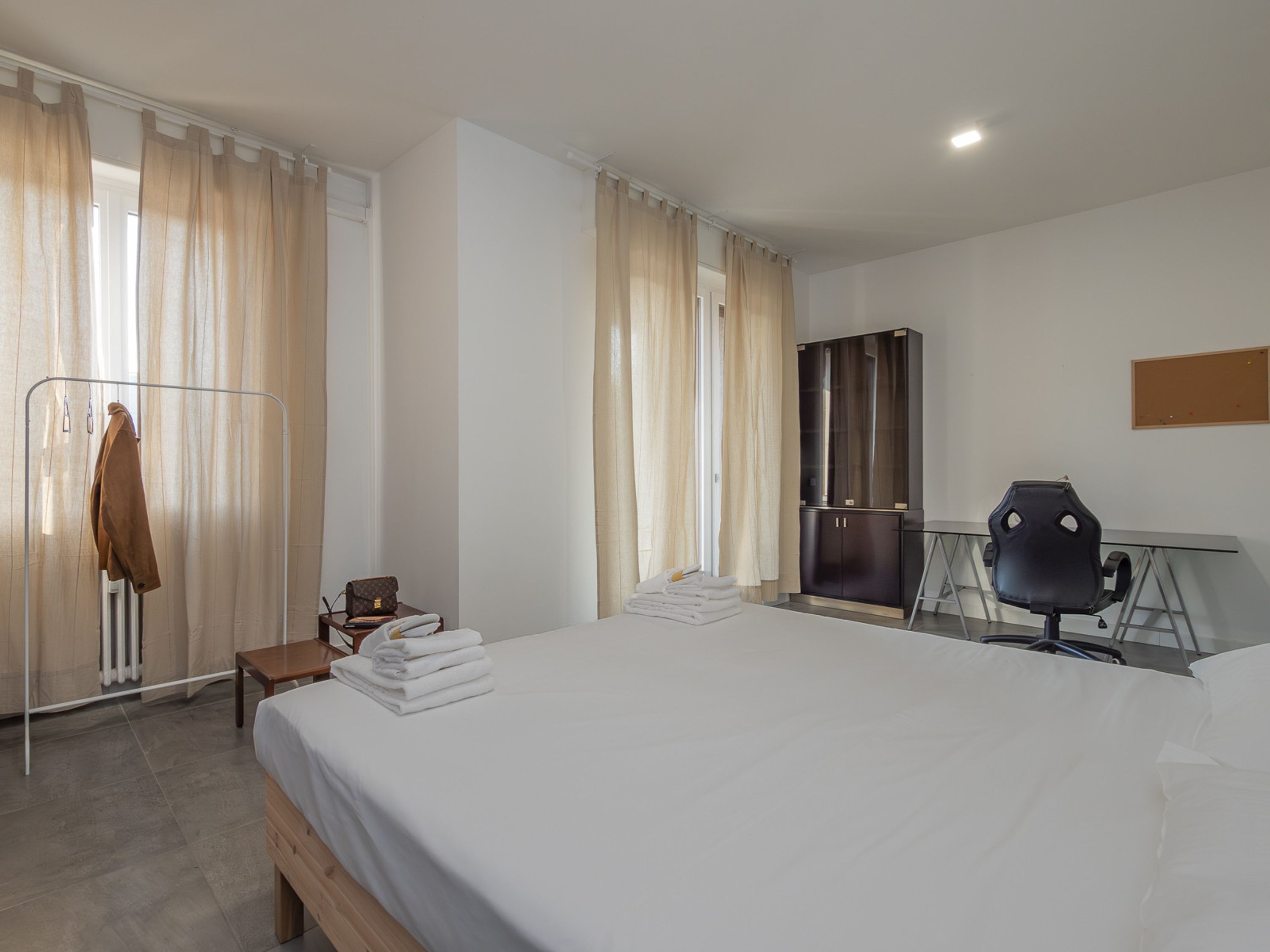 A hotel room with a large bed, a desk with a chair, a wardrobe, and a coat rack with a jacket.