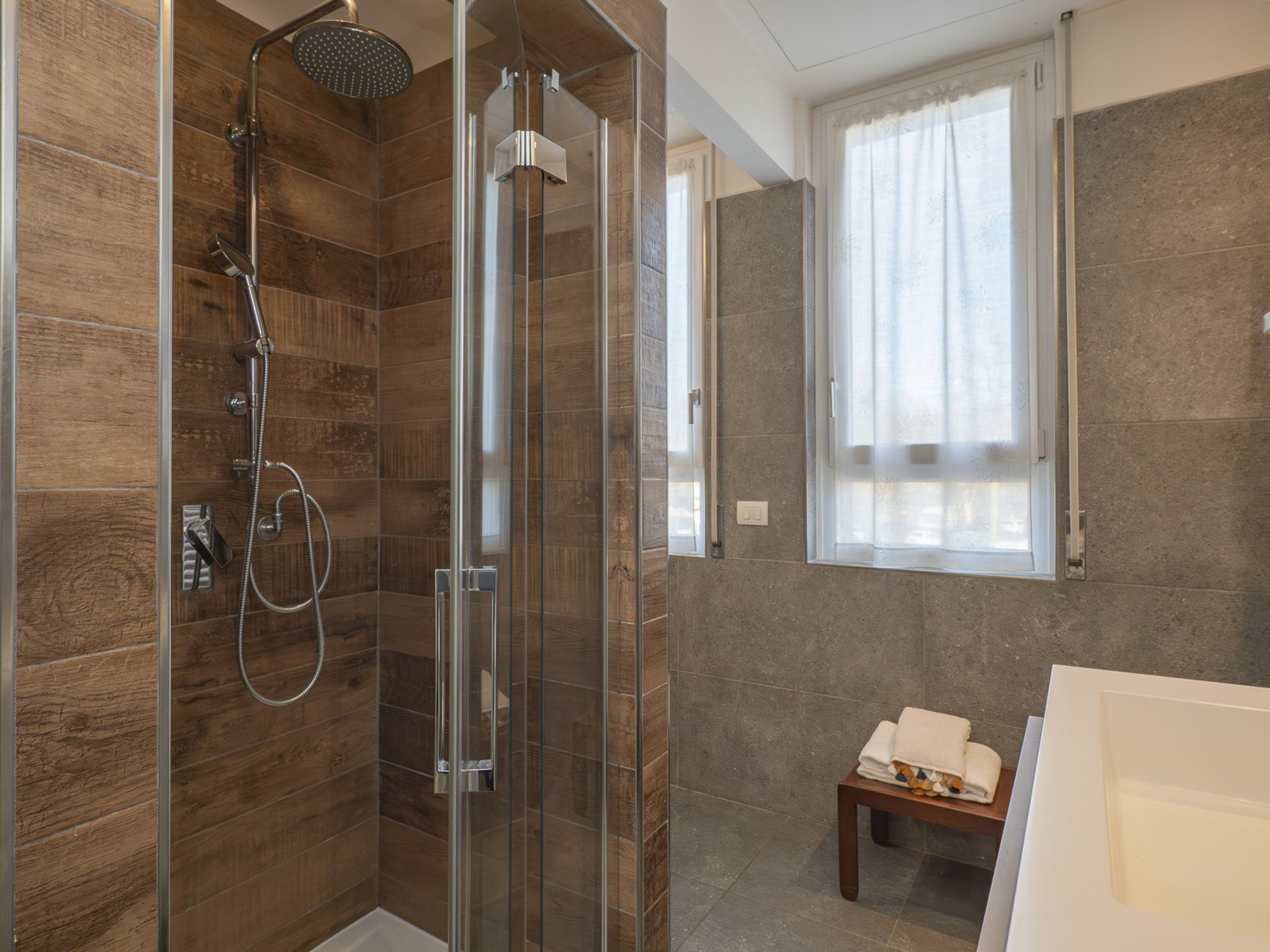 A modern bathroom with a glass-enclosed shower, a wooden-tiled shower area, a small wooden bench with towels, and a window with sheer curtains.
