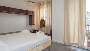 Moderni hotellihuone, jossa on puinen sänky, sisäänrakennettu vaatekaappi, ikkunassa verhot ja ikkunan edessä ruukkukasvi.