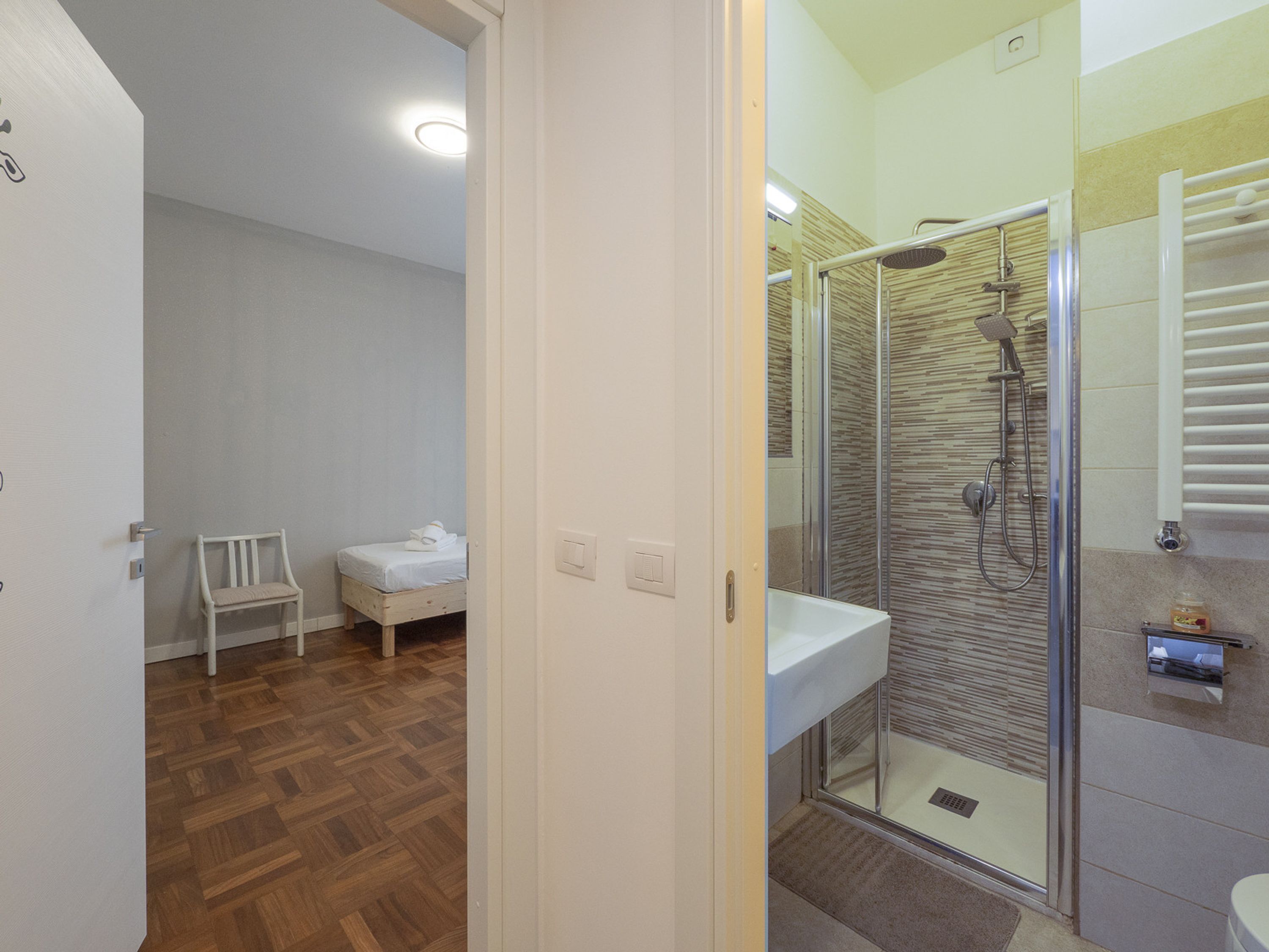 A modern bathroom with a glass shower enclosure, a small bedside table, and a chair.