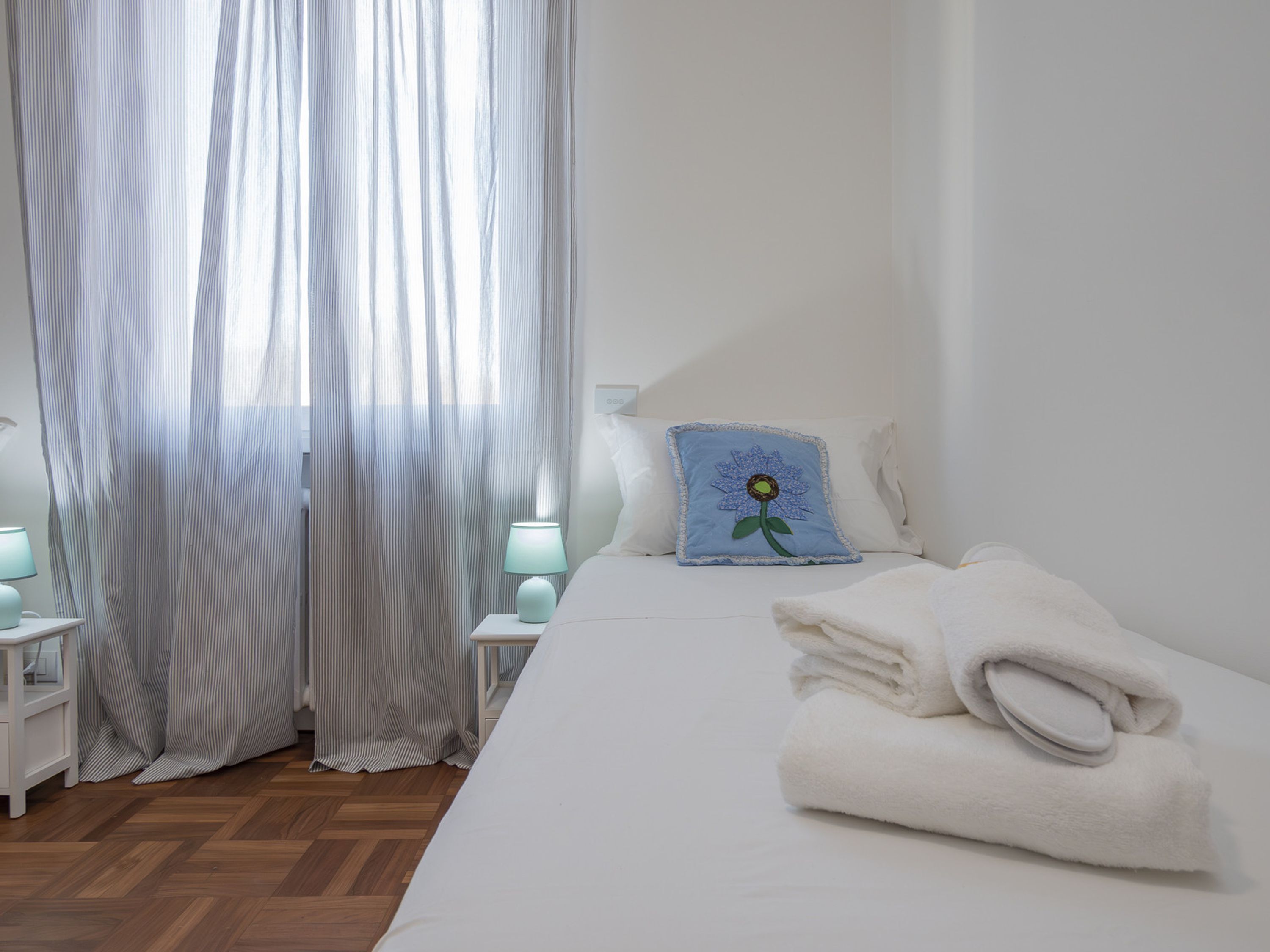 A hotel room with a neatly made bed, a pillow with a blue flower design, and a pair of folded towels on the bed.