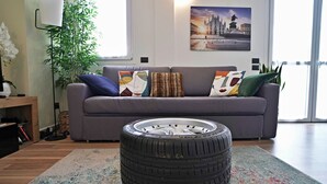 A living room with a grey sofa, colorful cushions, a large window, and a plant. - Palazzo Candiani - Modern Split-level 1BR Apartment Near Politecnico (Milan)