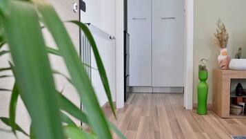 Un pasillo con piso de madera, una puerta blanca y un radiador. Hay una planta en primer plano y un mueble pequeño con un florero a la derecha.