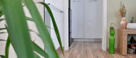 Um corredor com piso de madeira, porta branca e radiador. Há uma planta em primeiro plano e um pequeno armário com um vaso à direita.
