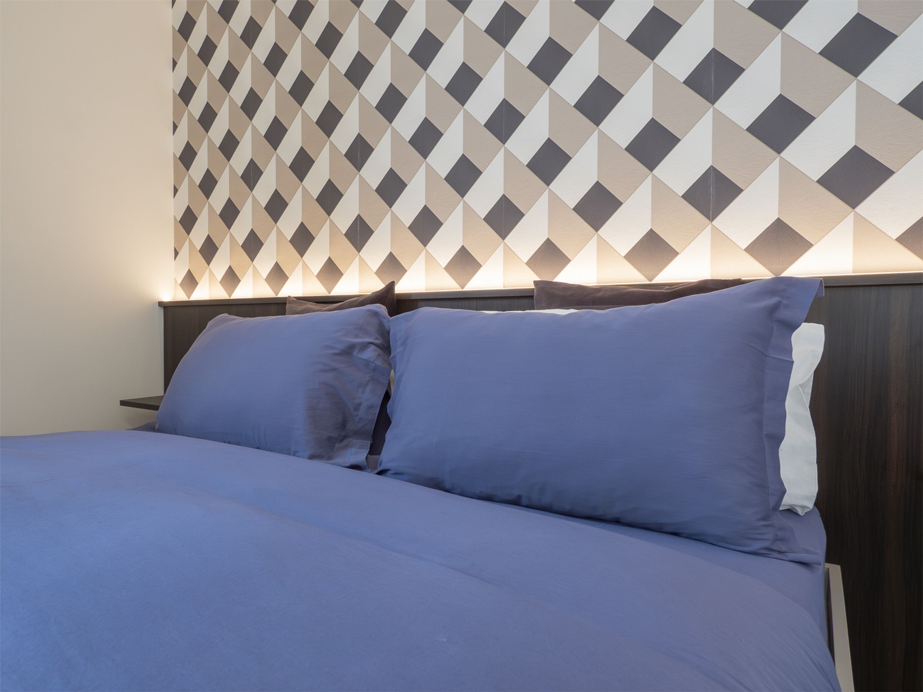 A bed with a blue mattress and pillows, a wooden headboard, and a wall with a geometric patterned wallpaper.