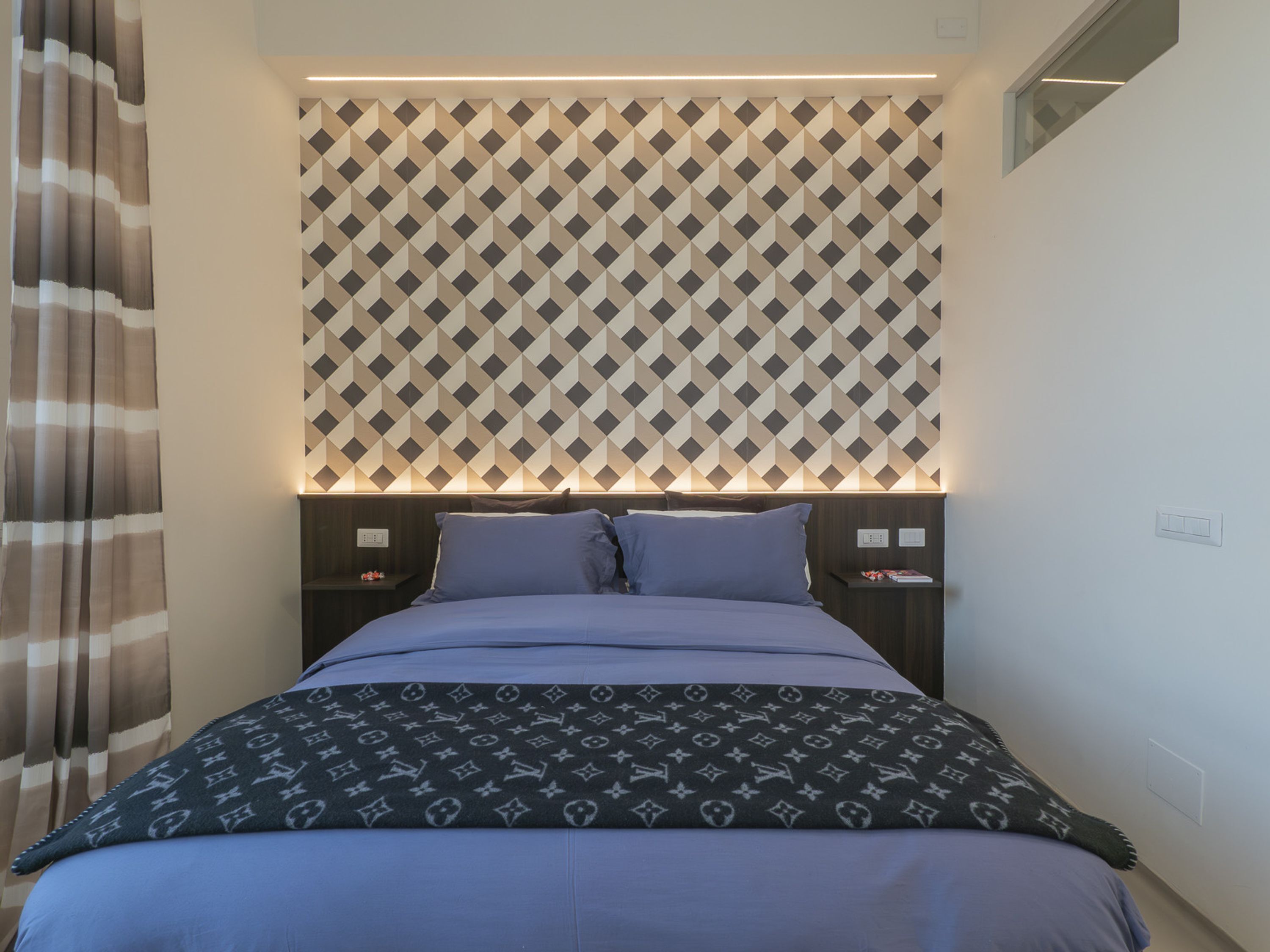 A neatly made bed with a patterned bedspread, a headboard with a geometric pattern, and bedside tables with lamps.