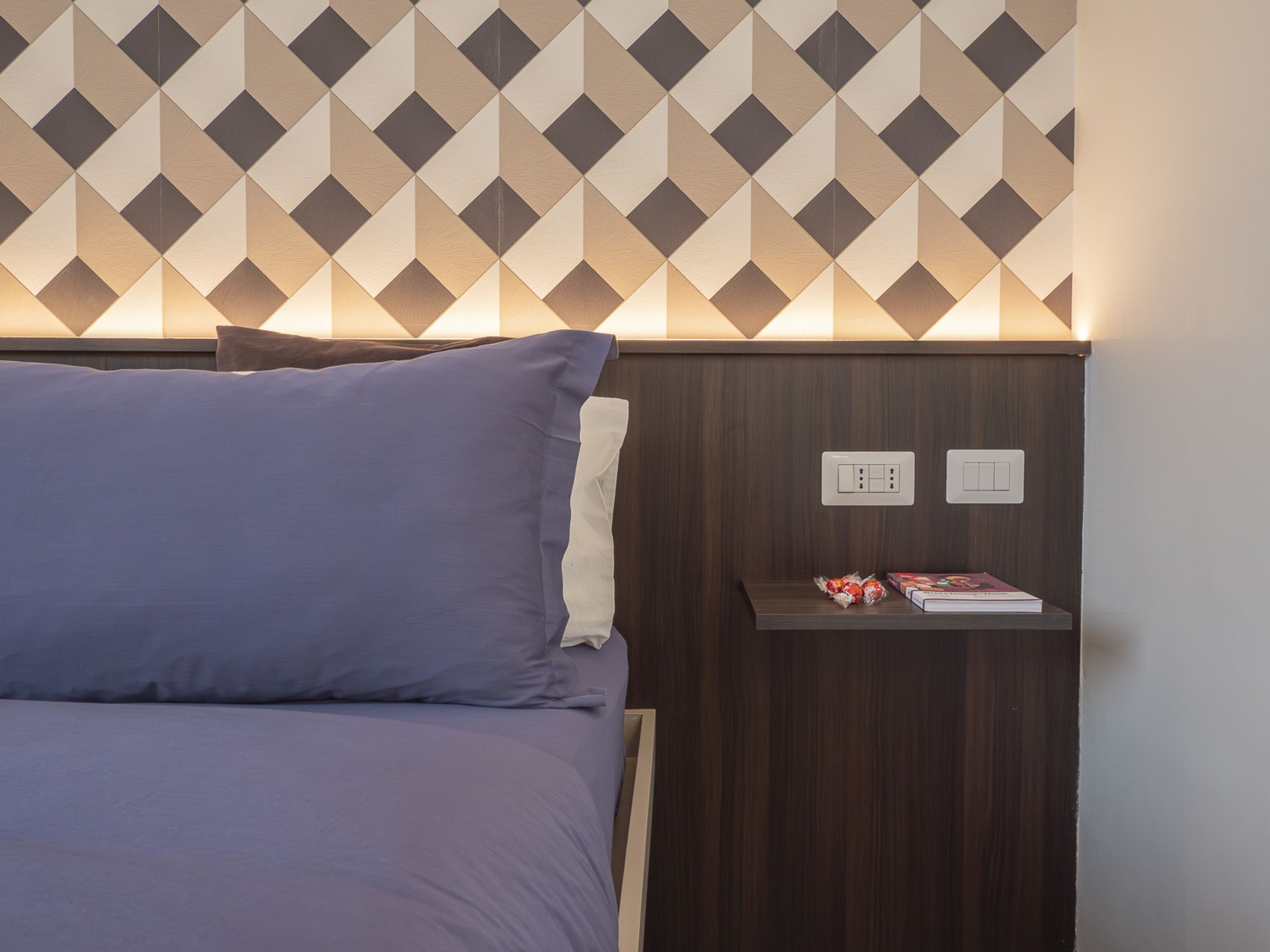 A neatly made bed with a blue blanket and pillows, a wooden headboard with a geometric pattern, and a bedside table with a book and a small decorative item.