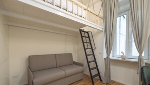 Een kamer met een houten vloer, een beige bank, een zwarte ladder, een raam met gordijnen en een klein plantje op de vensterbank.