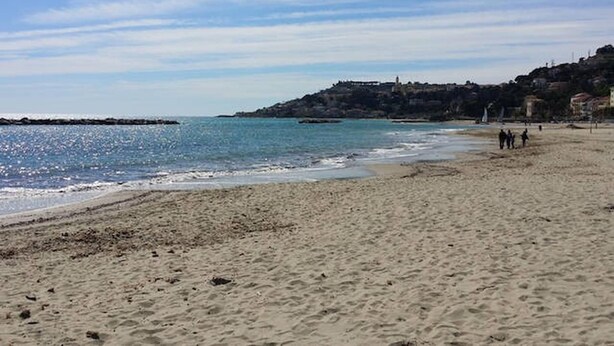 Une plage avec un promontoire rocheux au loin, une rive sablonneuse et quelques personnes qui se promènent.