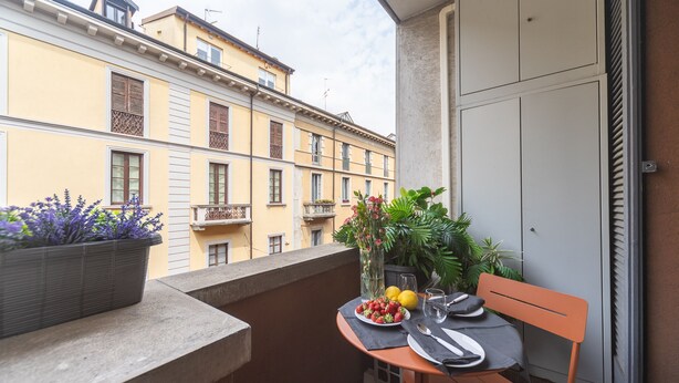 Un balcón con una mesa para dos, una planta en maceta y vistas de edificios cercanos.