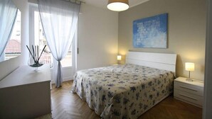 Een slaapkamer met een bed, bedlampen, een raam met gordijnen, een schilderij aan de muur en een nachtkastje met kaarsen.