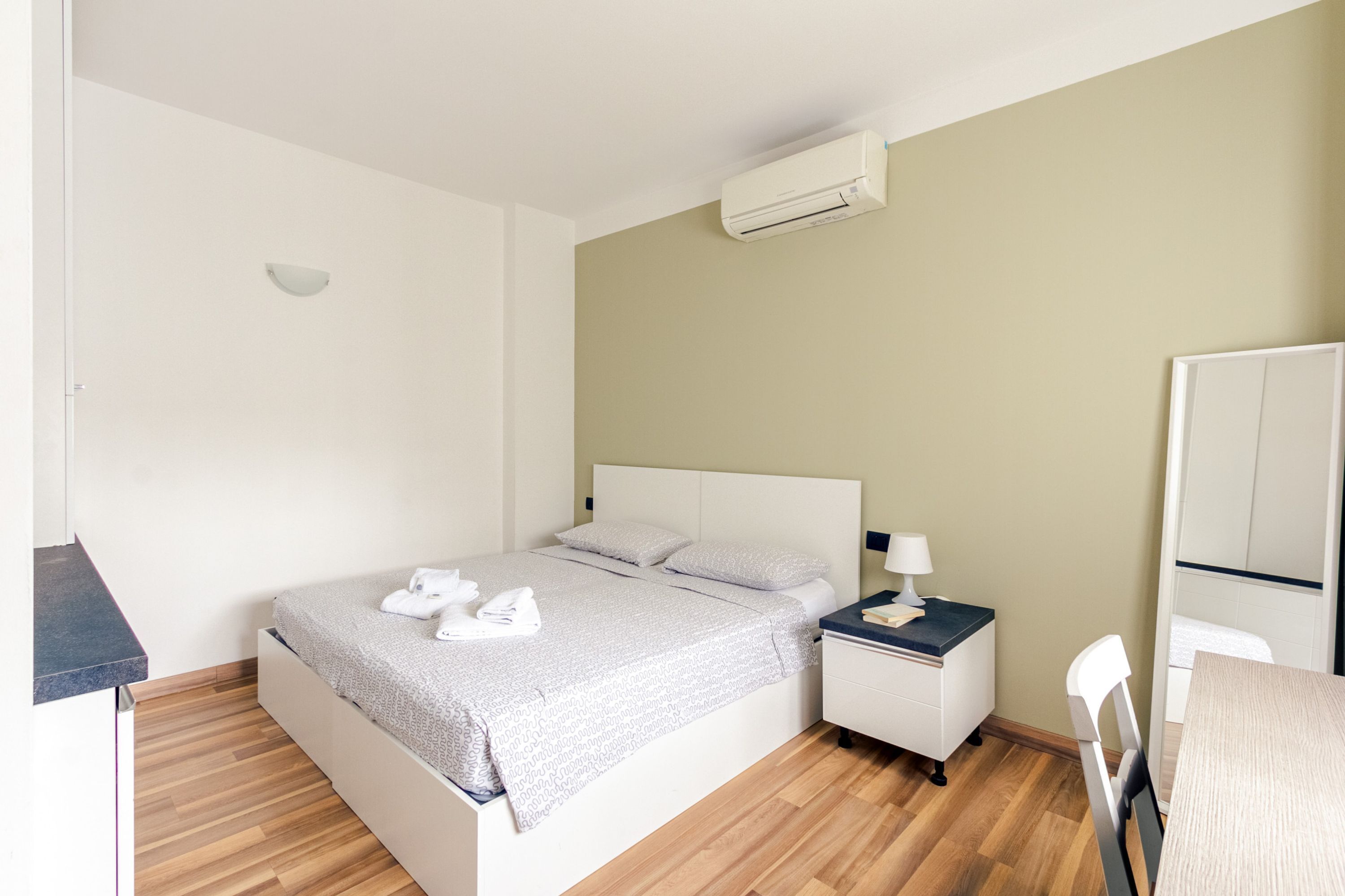 A bedroom with a bed, bedside table, lamp, and a mirror.