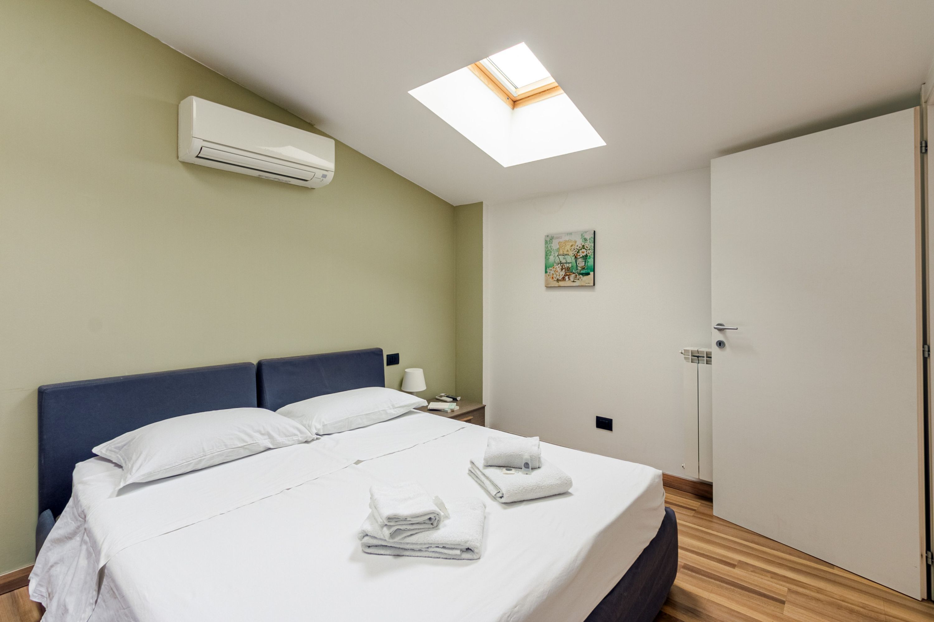 A hotel room with a bed, a skylight, an air conditioning unit, a nightstand, a painting, and a door.
