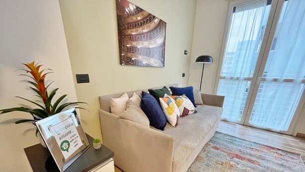 A modern living room with a beige sofa, colorful pillows, a framed welcome sign, a potted plant, and a large window with blinds. - Candiani Building - Modern 2BR Apartment Steps From Bovisa Politecnico (Milan)