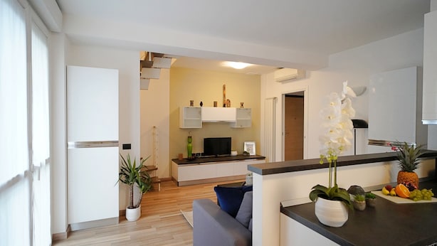 A modern kitchen with white cabinets, a built-in oven, and a central island with a flat-screen TV. There is a dining area with a gray sofa and a small table. - Candiani Building - Modern 2BR Apartment Steps From Bovisa Politecnico (Milan)