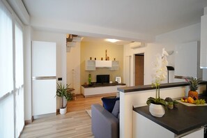 Een moderne keuken met witte kasten, een ingebouwde oven en een centraal eiland met een flatscreen televisie. Er is een zithoek met een grijze bank en een kleine tafel.