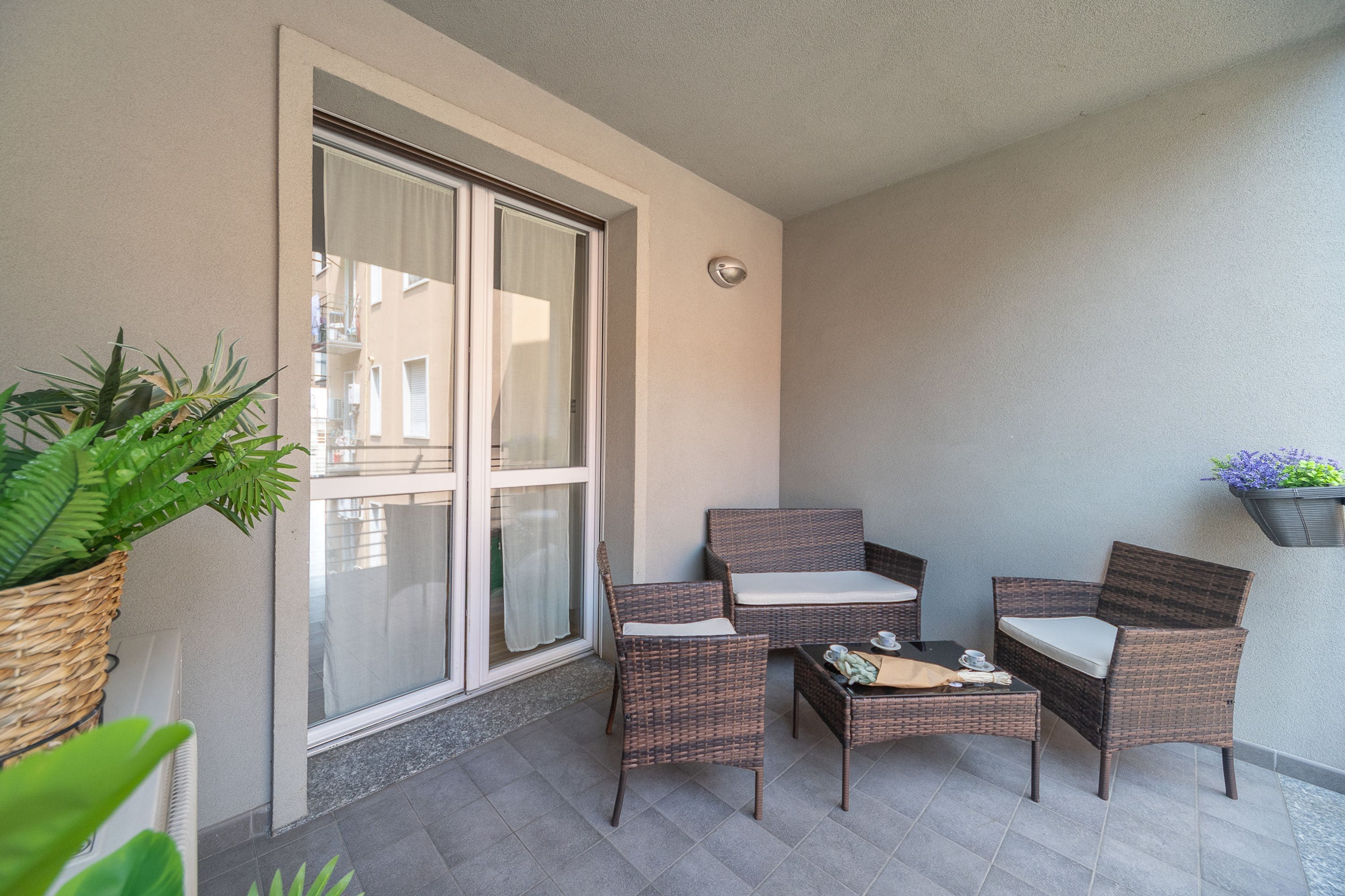 Ein Balkon mit Korbmöbeln, einem Blumentopf und Blick auf ein Gebäude durch eine Glastür.