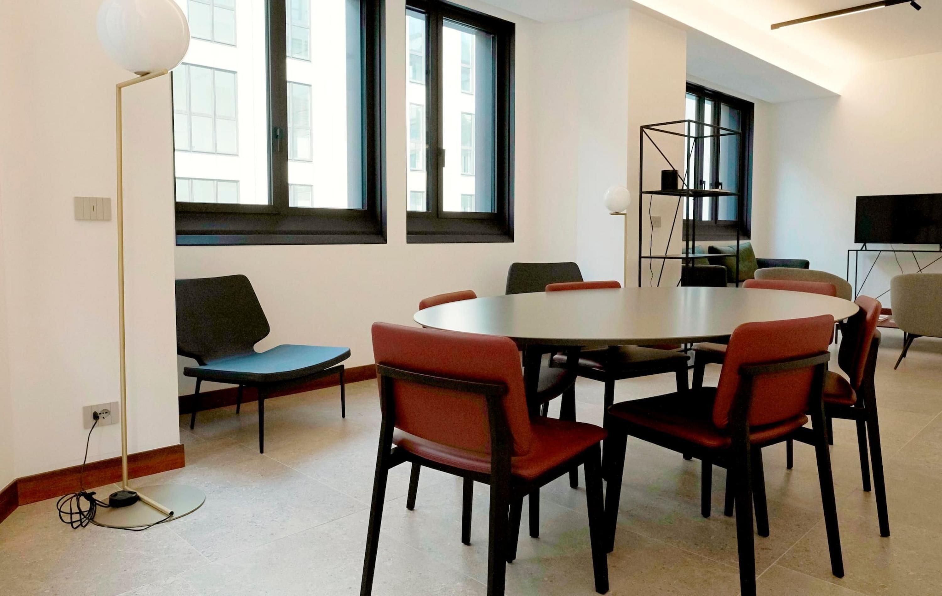 A conference room with a round table, six chairs, a floor lamp, and a TV on a stand.