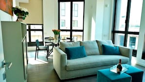 A modern living room with a grey sofa, two blue cushions, a glass coffee table with a bottle, a dining area with a table and chairs, and large windows with a city view. - S Babila Central Tower w Rooftop Terrace in San Babila (Milan)
