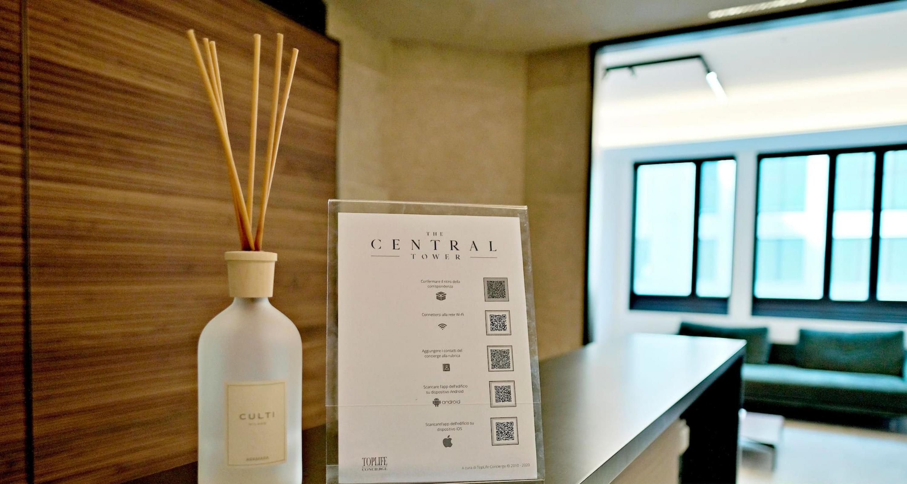 Un área de recepción de diseño moderno, con un revestimiento de madera, una mesa de recepción, una botella de velas Culti y un cartel con códigos QR.
