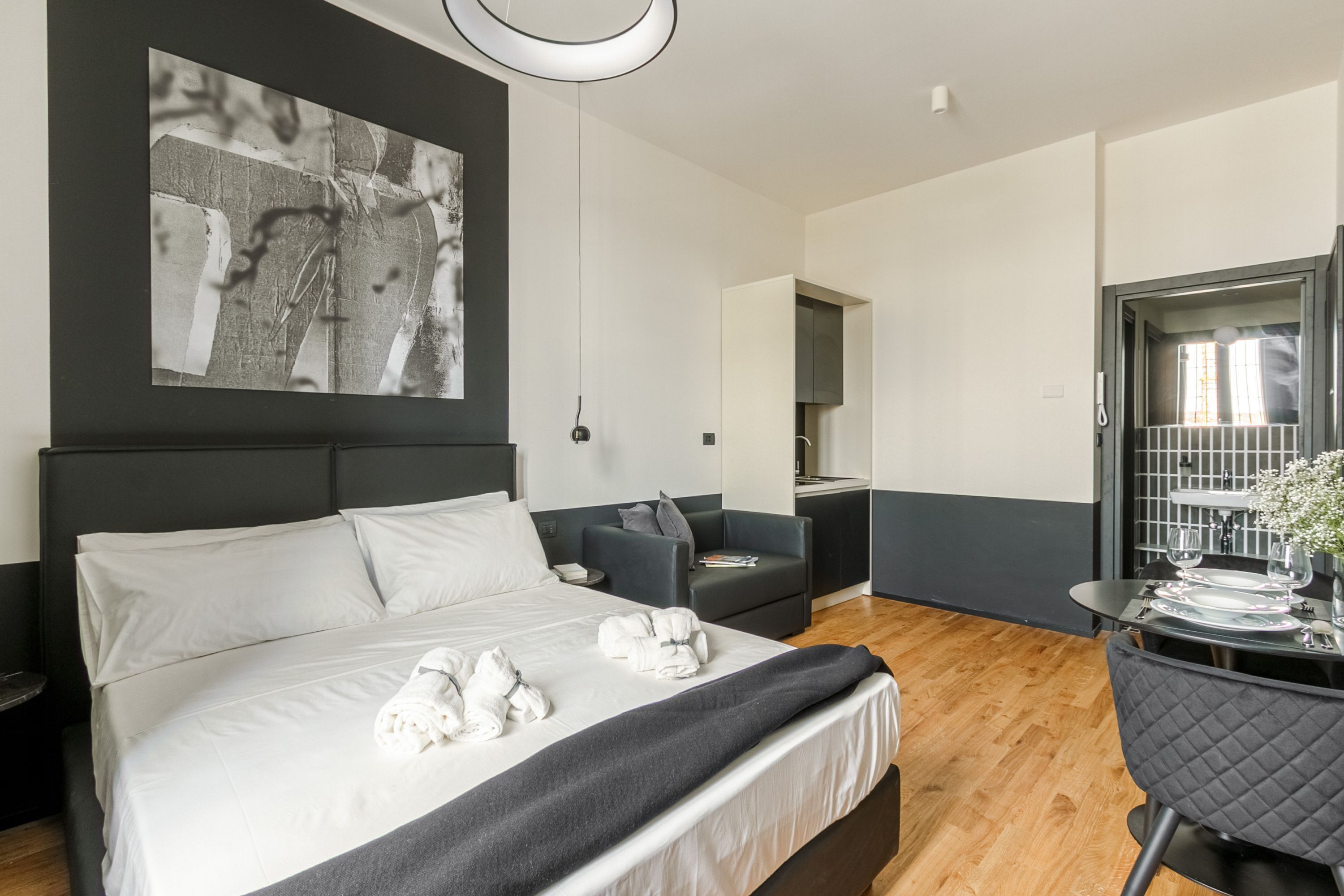 A modern hotel room with a large bed, a seating area with a sofa and armchair, a dining table set for two, and a large abstract painting on the wall.