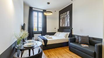 Quarto de hotel moderno com cama, sofá, mesa de jantar com pratos e copos, um vaso com flores e um quadro na parede.