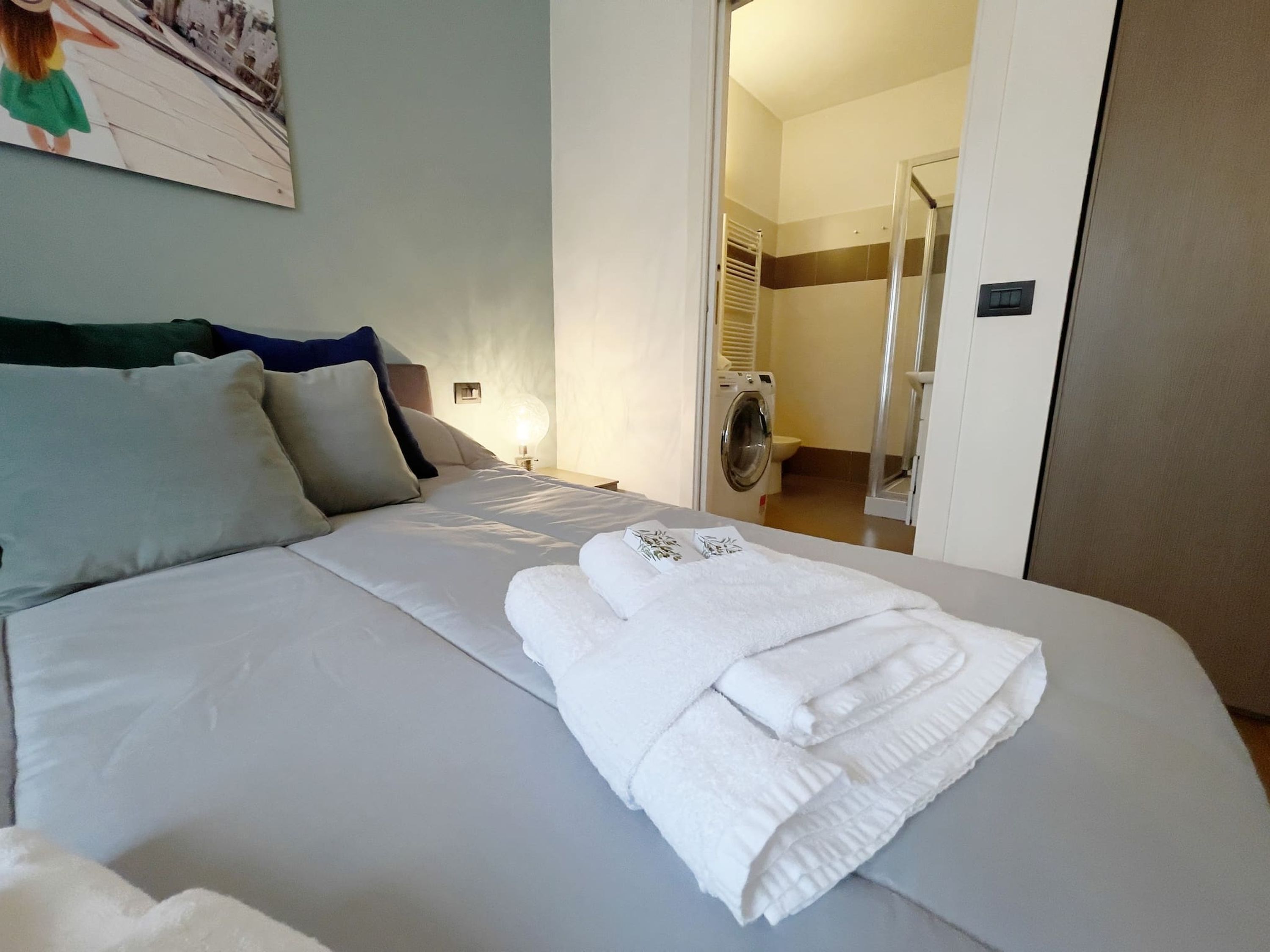 A neatly made bed with white linens and towels, a headboard, a bedside lamp, and a view into a bathroom with a washing machine.