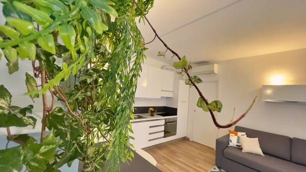 Una sala de estar moderna con un sofá gris y una alfombra estampada. Se ve una cocina con armarios blancos y una estufa al fondo.