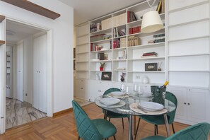Un coin repas avec une table en verre, des chaises à revêtement vert et une bibliothèque blanche remplie de livres et d’objets décoratifs.