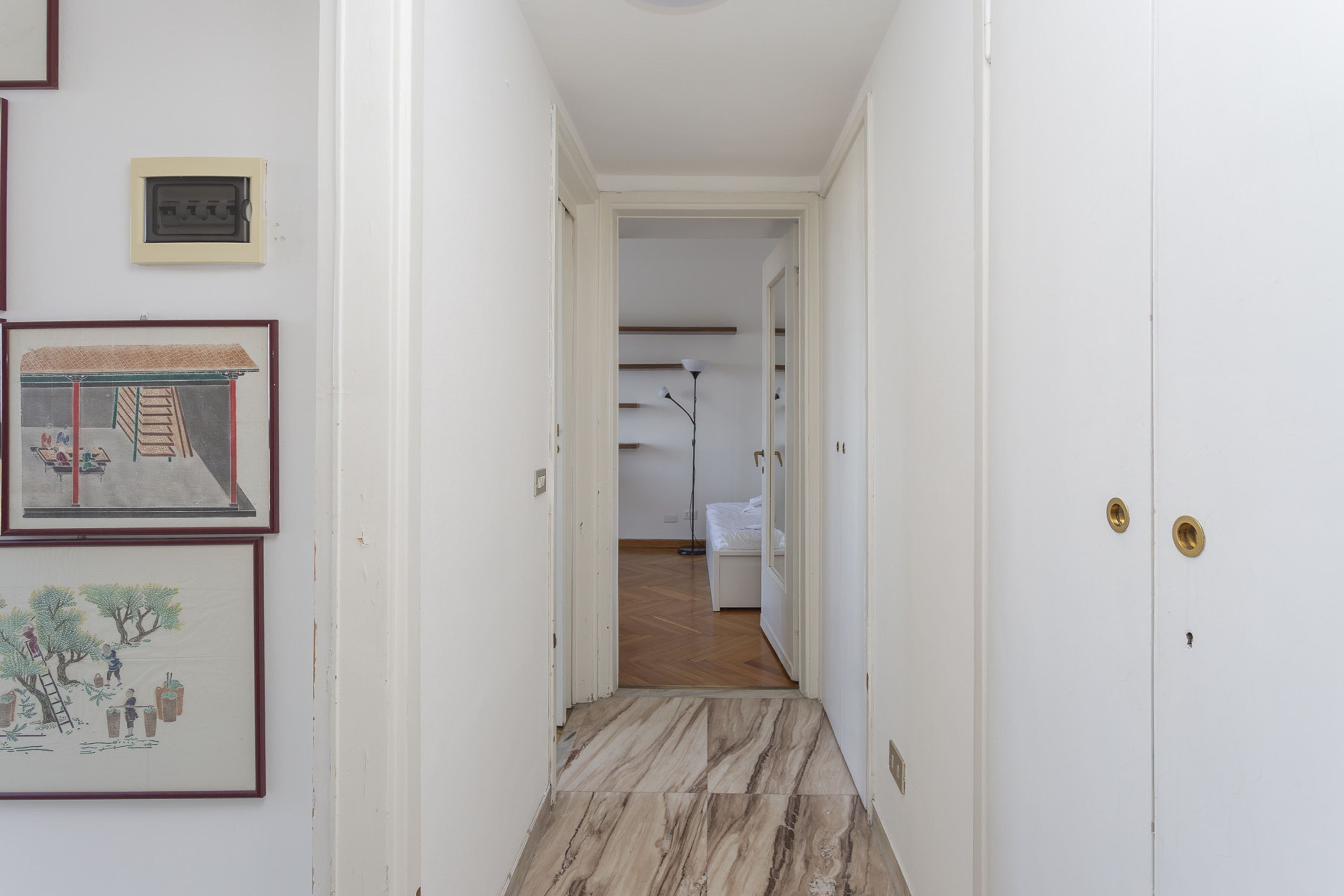 A narrow hallway with a wooden floor leading to a bedroom with a bed and a nightstand.