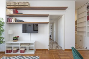Een moderne woonkamer met een flatscreen televisie aan de muur, houten boekenplanken, een witte console tafel met een lamp en een groene fauteuil.