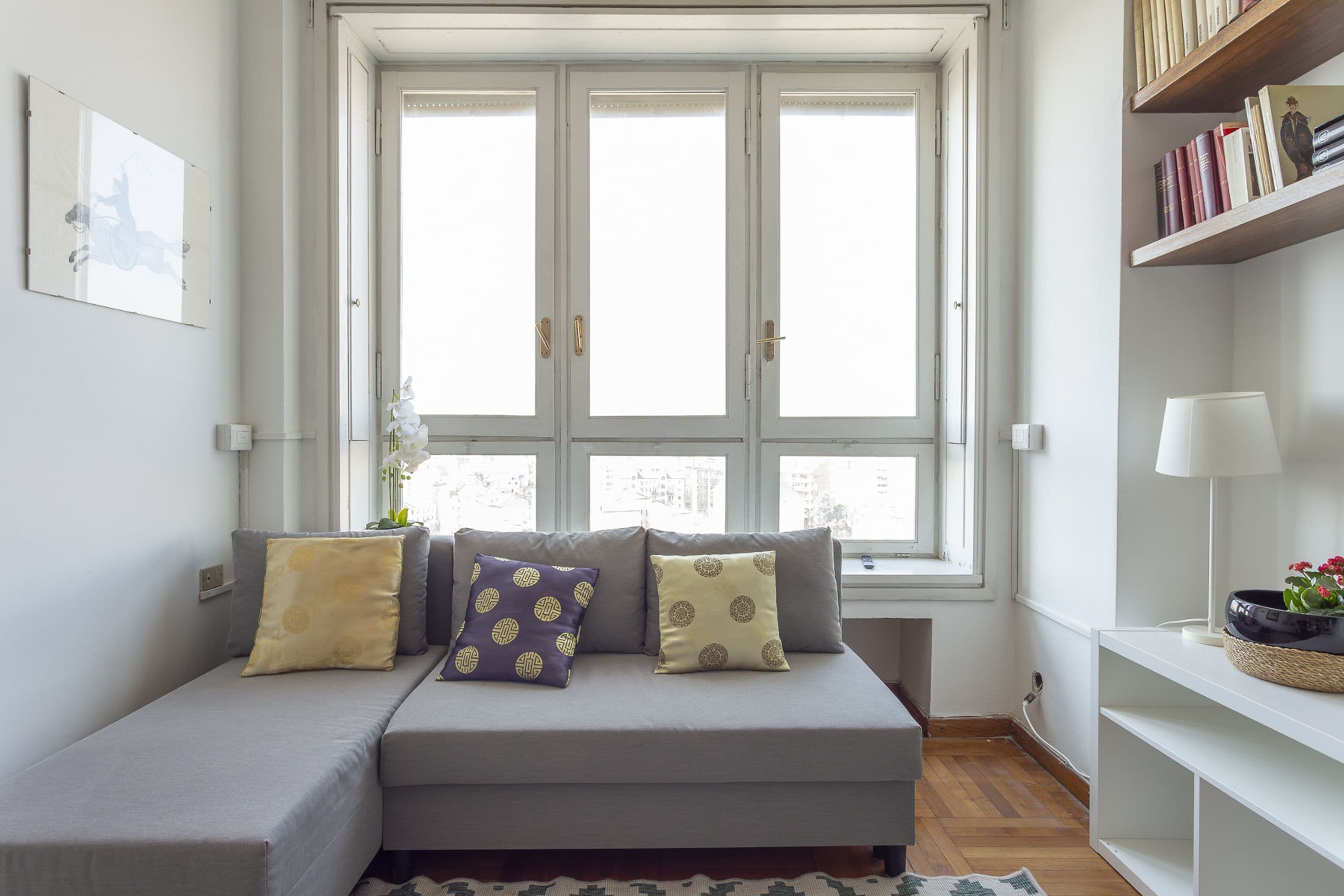 A modern living room with a grey sofa, two pillows, a white lamp, and a bookshelf.