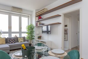 Ein modernes Wohnzimmer mit Sofa, Esstisch mit Stühlen, einem an der Wand montierten Fernseher und einem Bücherregal.