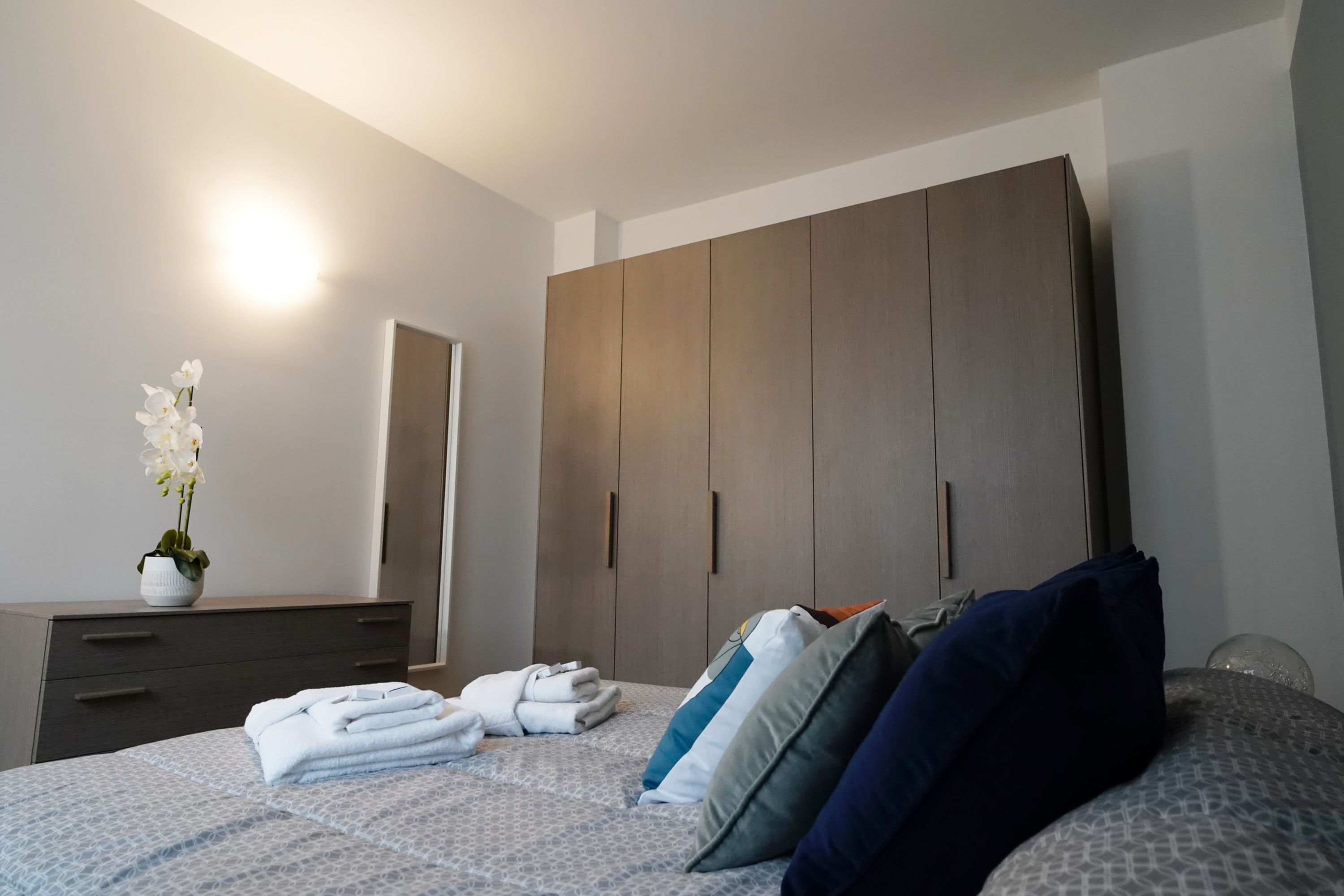 En pænt redt seng med et mønstret sengetæppe, et natbord med en vase med hvide blomster, og en stor garderobe med et spejl.