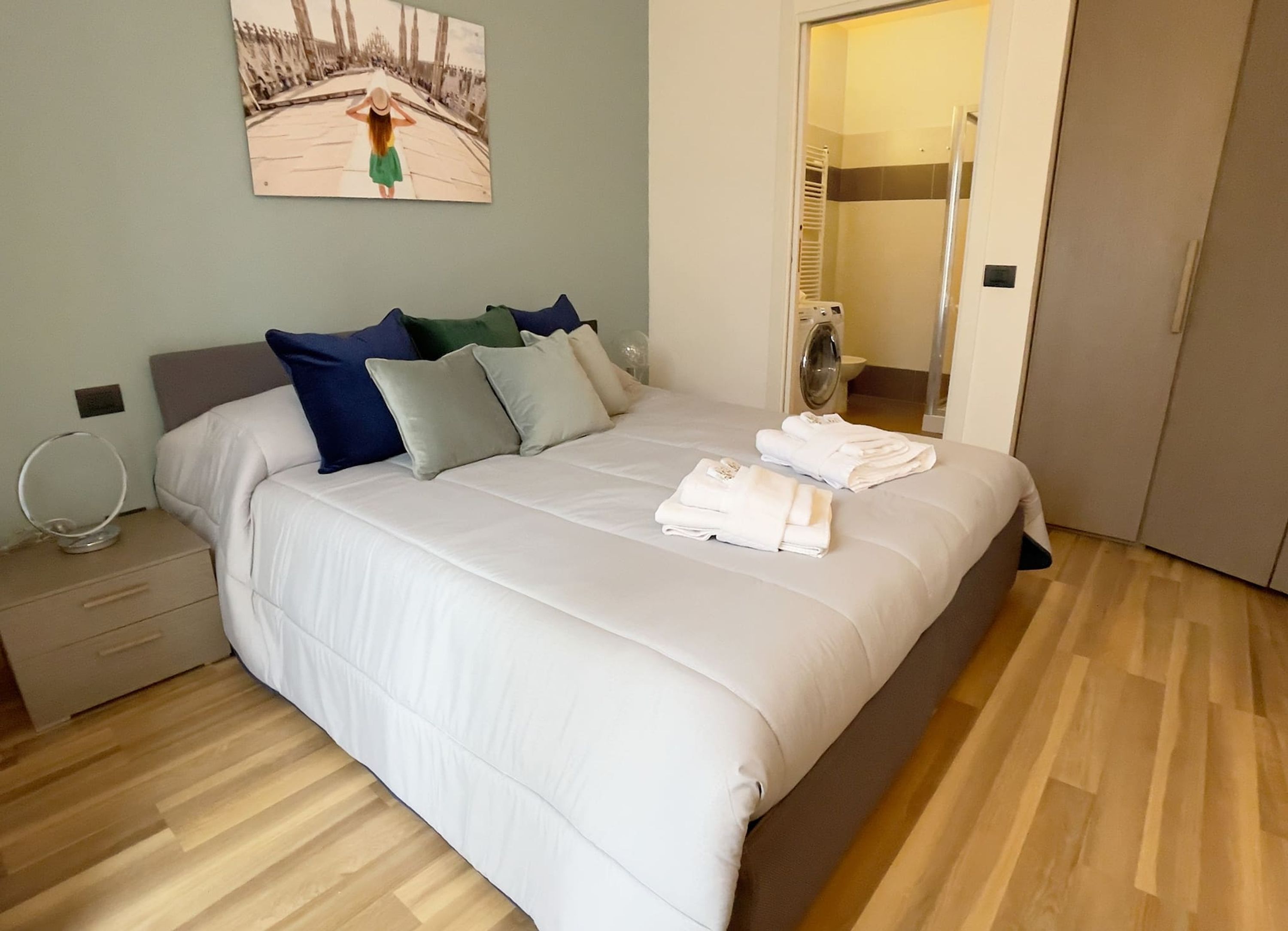 A neatly made bed with white linens and a few pillows, a bedside table with a lamp, and a painting on the wall.