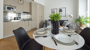 Et moderne køkken med mikrobølgeovn, vask og skabe. Der er et spisebord dækket med tallerkener, glas og bestik. En potteplante står på bordet, og et vindue giver naturligt lys.